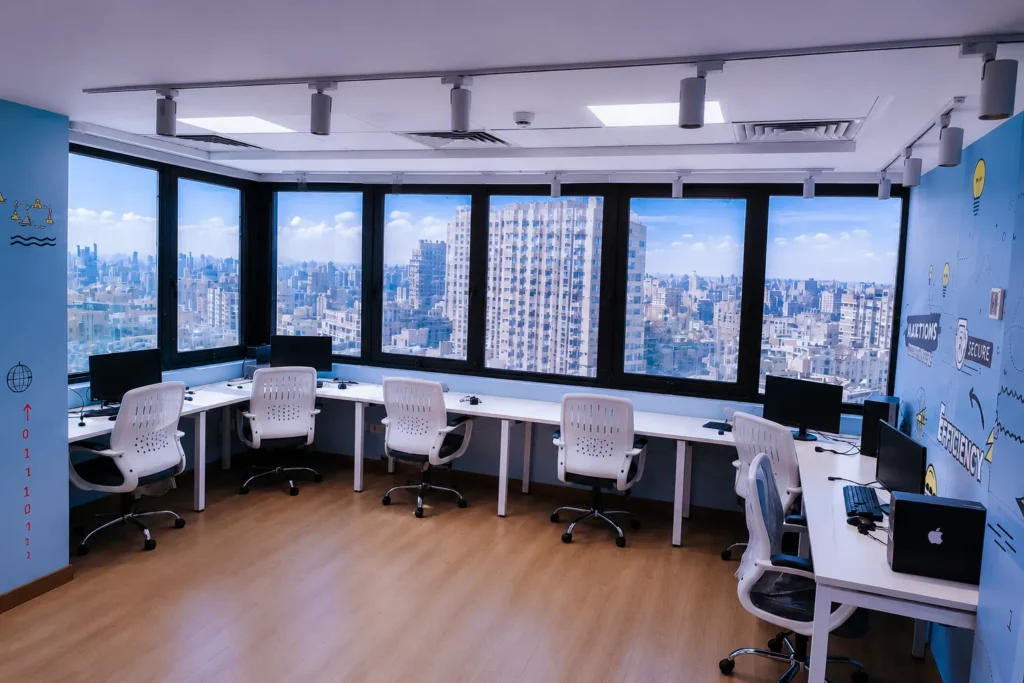 Bright open-plan office workspace with white desks, ergonomic chairs, computer stations, and expansive city views through large windows.
